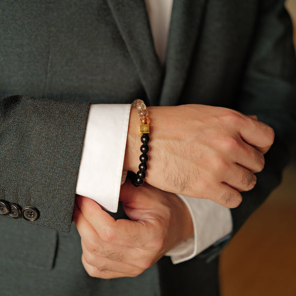 Close-up of a man adjusting his suit cuff, highlighting the glass-clear 8mm Black Gold Super Seven quartz filled with metallic bronze rutiles. Paired with high-gloss obsidian and a 14K gold PVD square signature bead, it fuels personal power and rapid manifestation.