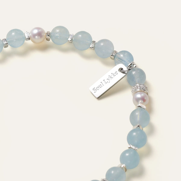 Close-up of the delicate aquamarine and Akoya pearl jewelry piece, highlighting the hypoallergenic 316L surgical steel signature tag finished in premium white gold PVD and 925 sterling silver pavé rondelles.