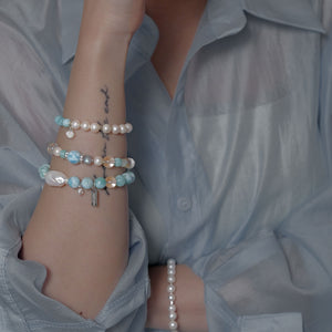 Close-up of a sophisticated demi-fine jewelry stack, highlighting the unique organic silhouette of a 20mm East Asian Baroque Pearl, ice-clear Brazilian Quartz, and the vivid sky blue tortoise-shell patterns of natural Larimar beads.