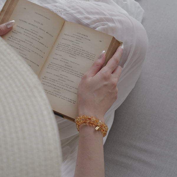 Close-up of a model holding an open book while wearing layered 6mm and 8mm Soul Lykke Citrine bracelets, capturing the dancing brilliance and varying bead scales of authentic honey-gold crystals paired with 24k gold-plated accents.