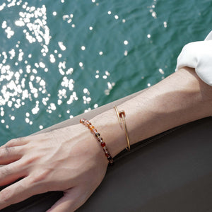 Model styling a 6mm Black Gold Super Seven crystal bracelet with a gold bangle against a sparkling ocean, showing a cool architectural edge.