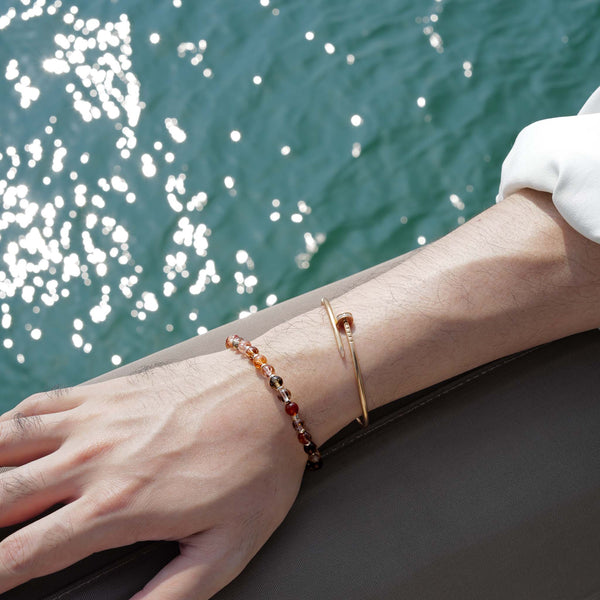 Model styling a 6mm Black Gold Super Seven crystal bracelet with a gold bangle against a sparkling ocean, showing a cool architectural edge.