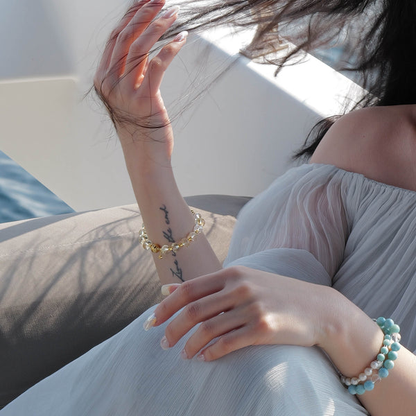 Model on a yacht styling a faceted AAA Citrine and Clear Quartz bracelet on one wrist, paired with a layered Larimar and Pearl stack on the other.