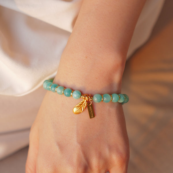 Close-up of the 7mm icy amazonite bracelet on a woman's wrist, revealing the stones' gel-like translucence in natural sunlight. Accented by a beautifully sculpted 24K gold-plated feather charm and hypoallergenic signature tag.