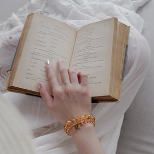 A peaceful lifestyle moment showing a woman reading a vintage book, wearing a beautifully layered stack of 6mm and 8mm AAA Brazil Citrine gemstone bracelets that symbolize mental clarity and quiet refinement.