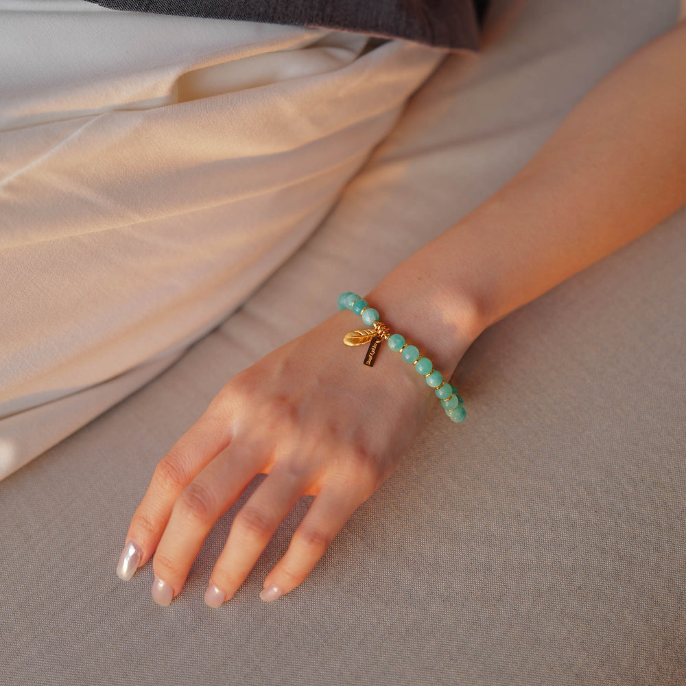 The Tide & Feather bracelet by Soul Lykke, featuring 7mm natural AAA Mozambique icy amazonite beads and a 24K gold-plated sculpted feather charm on a seamless neutral background. This is a natural gemstone and healing crystal bracelet by Soul Lykke.