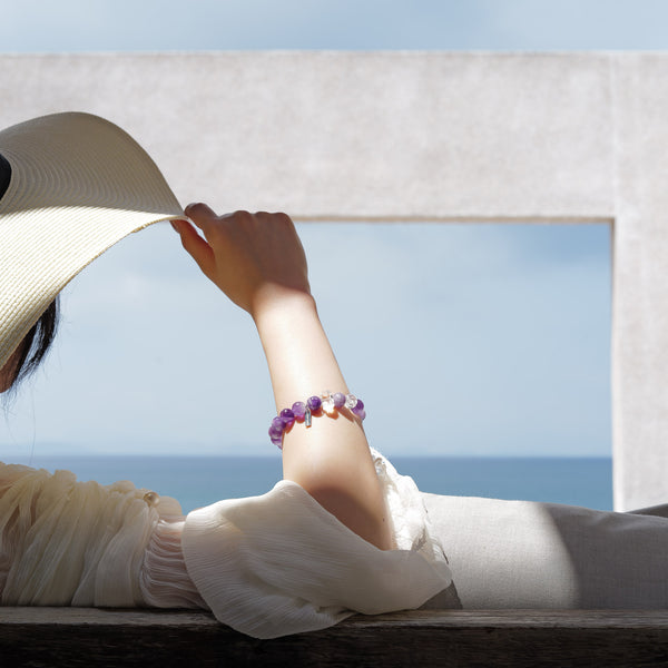 A woman relaxing by the ocean wearing a 10.5mm Dream Chevron Amethyst healing bracelet. Metaphysically crafted to activate the Third Eye and Crown Chakras, the sacred geometry of the stones filters mental clutter and shields against psychic overload.